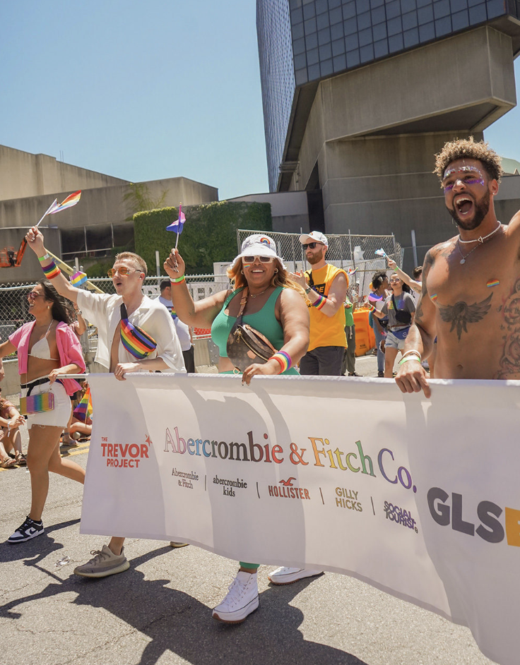 Individuals marching in a pride parade waving flags and holding a banner reading "Abercrombie & Fitch Co." as well as "The Trevor Project."