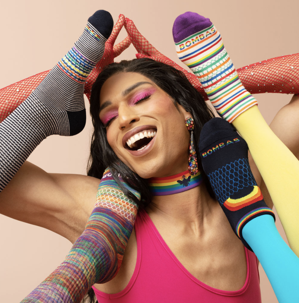 A person with a rainbow choker, pink eyeshadow, and red fishnet gloves on, posing between four different legs wearing various Bombas brand rainbow socks.