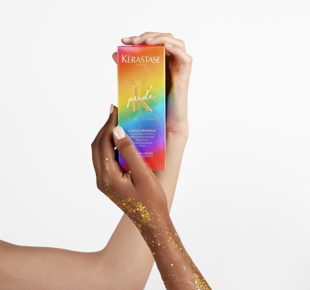 Hands holding and displaying a box containing Kerastase limited edition hair oil with PRIDE-inspired rainbow packaging in front of a white background.
